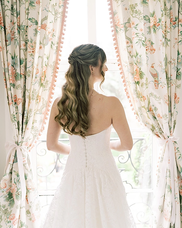 Bridal portrait of a bride looking out window from behind in a strapless lace gown, loose curls half-up, in bright balcony light with greenery
