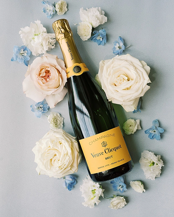 Champagne bottle wedding champagne flat lay with white and peach roses and blue blossoms arranged on a light gray surface