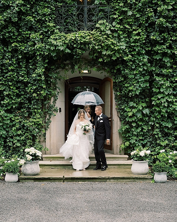 Bride walking out under a clear umbrella, holding a bouquet and veil, stepping from an ivy-covered stone doorway down to the gravel drive