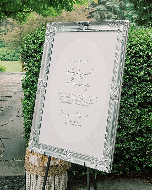 Wedding signage unplugged ceremony sign in an ornate silver frame on an easel, with programs in a basket along a stone garden walkway