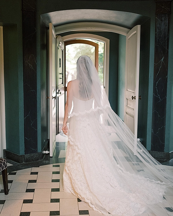 Bride portrait from behind with long cathedral veil and lace train, walking through an arched doorway into a teal-walled hallway with tile floor