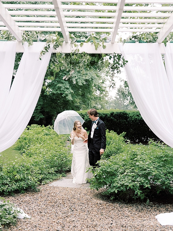 Couple portrait of bride and groom under umbrella, bride laughing in strapless dress beside tuxedoed groom on a garden path by a white pergola