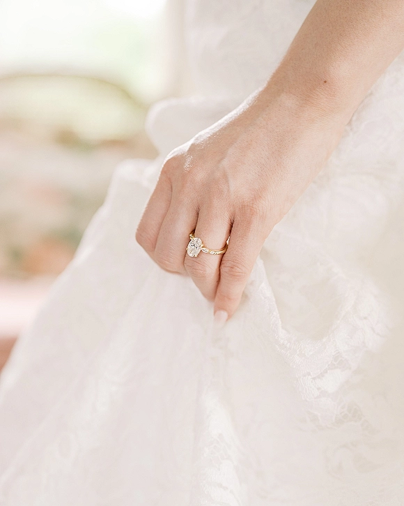 Engagement ring with an oval diamond on a thin gold band, shown on manicured hand holding lace bridal gown against a soft blur background