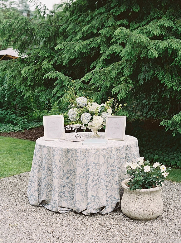 Wedding guest book table with hydrangea centerpiece, framed welcome signs, and vintage telephone on blue toile cloth in a garden setting
