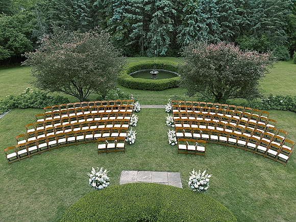 Ceremony setup with outdoor ceremony seating of wood chairs and white cushions, floral aisle markers leading to a garden fountain on a manicured lawn