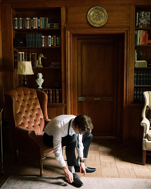 Groom getting ready, tying black leather oxford shoes while seated in a tufted armchair in a wood-paneled library with bookshelves