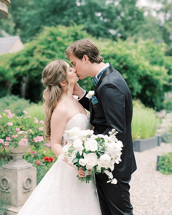 Wedding kiss as bride in strapless lace dress holds a white rose bouquet, embracing groom in black tuxedo by a stone garden balustrade