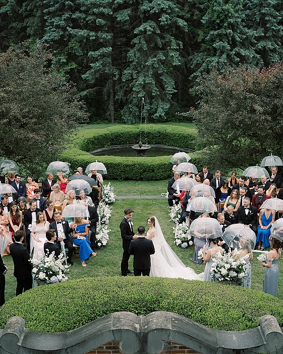 Outdoor wedding ceremony in a rainy wedding ceremony, guests under clear umbrellas along floral aisle on a manicured garden lawn with fountain