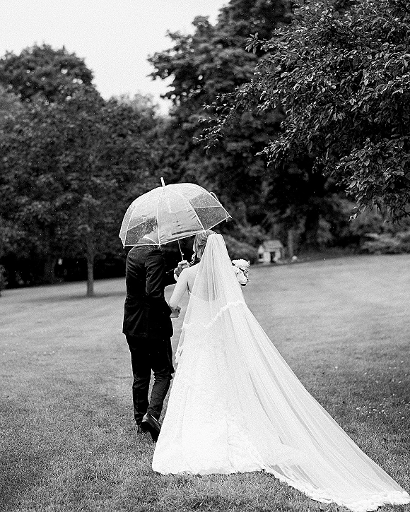 Wedding couple portrait of bride and groom walking away under a clear umbrella, her long veil and dress train trailing on a lawn with trees
