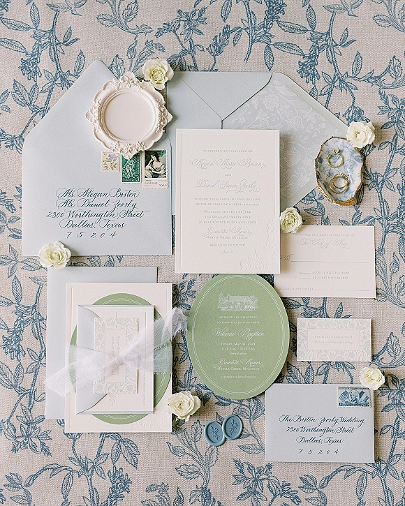Wedding invitation suite with blue and white invitations, wax seals, vintage stamps, ribbon, rings, and white roses on blue floral fabric