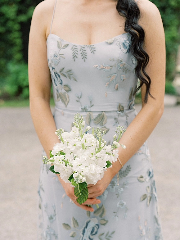 Bridesmaid portrait holding bouquet of white flowers and greenery in front, wearing a light blue floral spaghetti-strap dress on a garden path