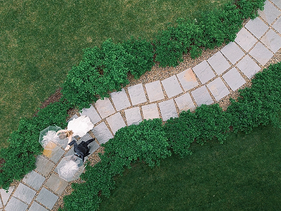 Couple portrait from above of bride and groom walking with clear umbrellas, veil and dress train flowing along a stone walkway bordered by lawn