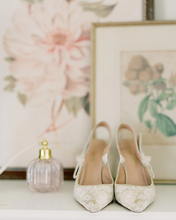 Wedding shoes in a bridal shoe detail flatlay, white pointed-toe slingback heels in floral brocade beside perfume on a white tabletop with framed botanical art