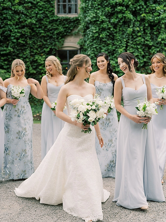 Bride with bridesmaids laughing in blue dresses, holding bouquets beside an ivy-covered wall and window on a garden gravel path
