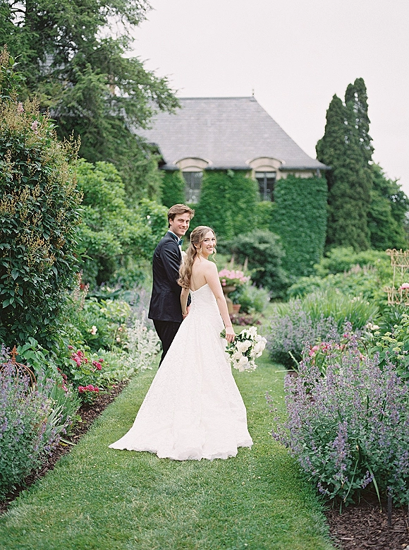 Couple portrait of bride and groom holding hands, walking a garden path as she looks back, holding a white rose bouquet in a tuxedo and strapless dress train