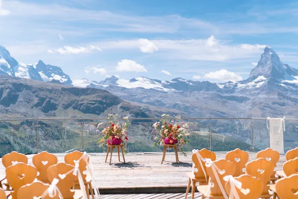 Ceremony setup with wooden chairs and white ribbons, colorful florals on stools beside a glass railing overlooking snowcapped peaks