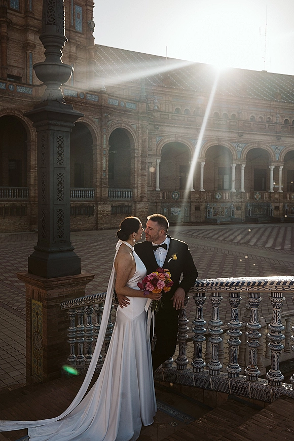 Couple portrait of bride and groom embrace, bride holding bouquet with long veil in ornate courtyard, sun flare by arched brick building