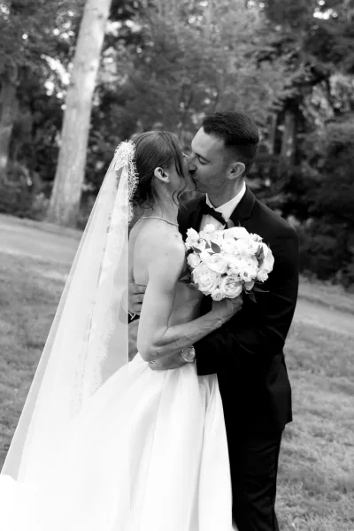 Wedding kiss portrait of a bride and groom under garden trees, her lace-trimmed veil framing their embrace on the grass lawn.