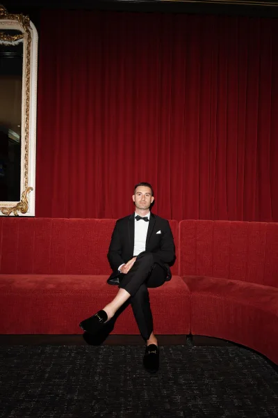 Groom portrait seated on a red sofa before a gilded mirror frame, wearing a bow tie, against a red velvet curtain and dark carpet.