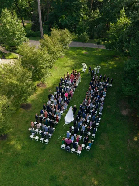 Ceremony moment during the wedding processional as the couple walks past aisle petals along a tree-lined lawn and garden path.