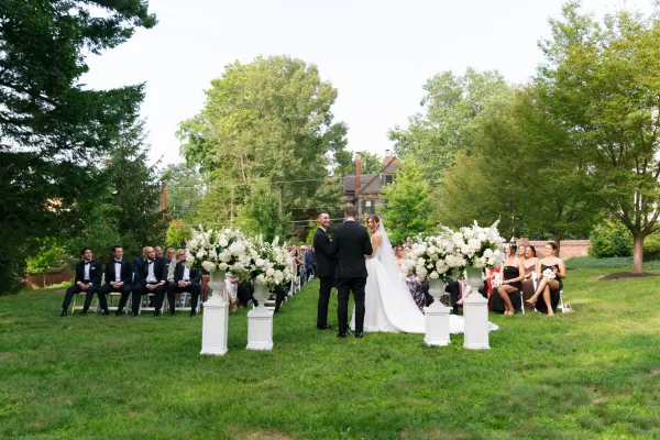 Ceremony moment at an outdoor wedding ceremony as the bride and groom stand, framed by white florals on a tree-lined lawn.