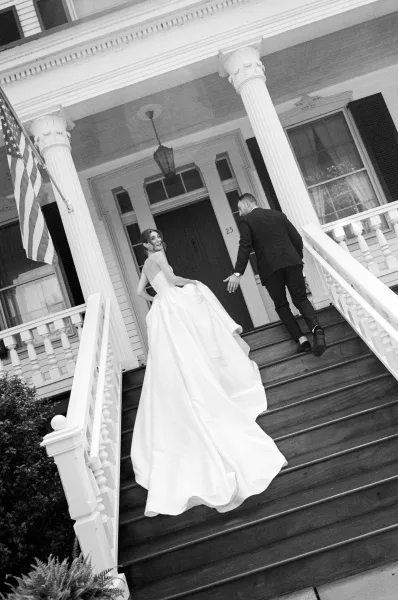 Couple portrait with the bride and groom walking up a columned front porch staircase, her long train sweeping the steps as he follows.