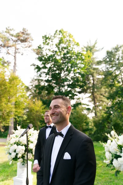 Groom portrait, a groom waiting at the altar in a classic bow tie, set against garden trees, grass lawn, and sky.