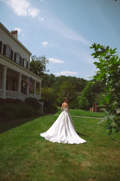 Bridal portrait of a bride walking away, her dress train trailing across a grass lawn toward a historic house with porch columns and trees.