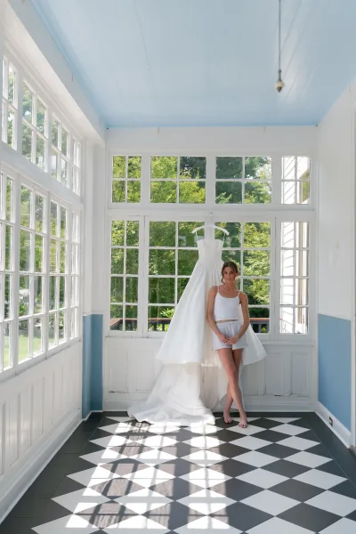 Bride getting ready with her dress on a hanger, standing by large windows on a checkerboard floor with garden trees beyond.