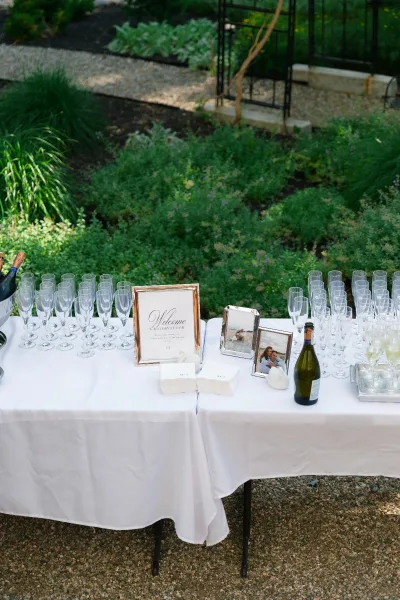 Champagne bar setup as a welcome drink station with a framed welcome sign, set by garden greenery and a metal arbor on gravel.