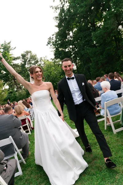 Ceremony recessional during a just married exit, bride and groom walk the aisle as she lifts her bouquet, with garden trees and seated guests behind.