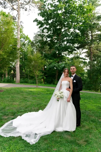 Couple portrait of a bride and groom standing on a garden pathway beneath leafy trees, the bride holding a classic white bouquet.