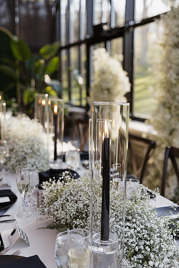 Reception tablescape with baby’s breath centerpiece garland, black taper candles and glass hurricanes on white linens by sunlit windows