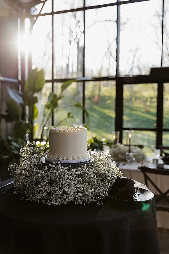Wedding cake with smooth buttercream on a stand, accented by baby's breath, taper candles, and greenery near sunlit industrial windows