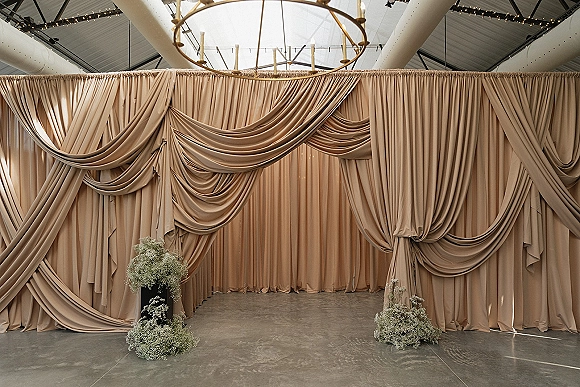 Wedding backdrop with draped wedding backdrop fabric, chandelier and string lights over floral pedestals in an industrial venue with skylight