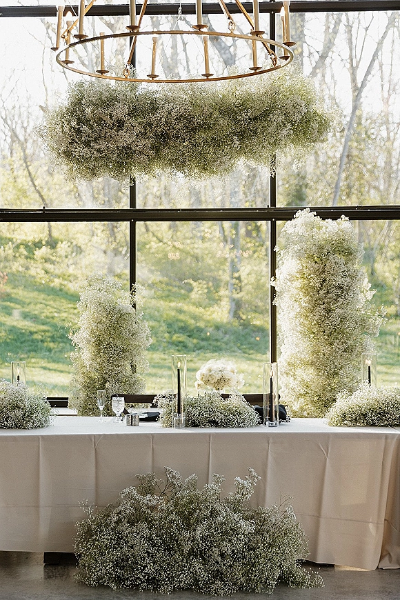 Reception sweetheart table with baby's breath florals, taper candles in glass cylinders, and black napkins beneath chandelier by window walls
