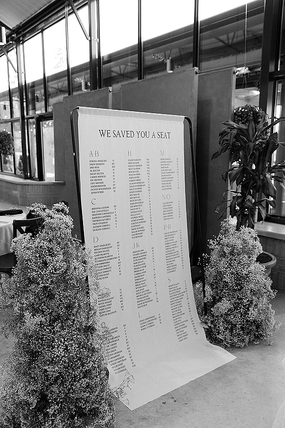 Wedding seating chart displayed on a black metal stand with printed list and baby’s breath accent in an industrial glass-walled venue