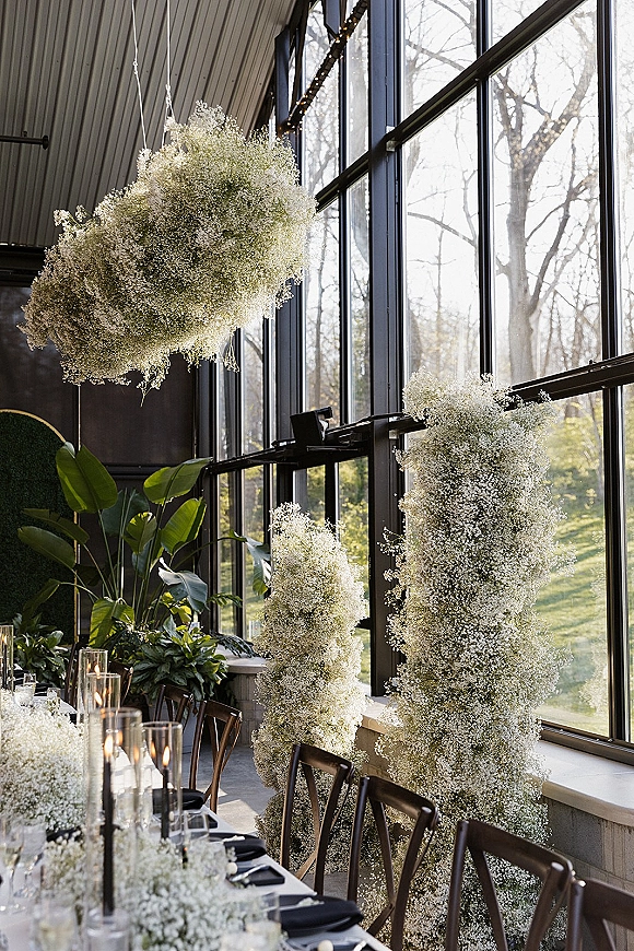 Reception tablescape with baby’s breath accents, taper and tall glass cylinder candles beneath a hanging floral cloud by industrial windows