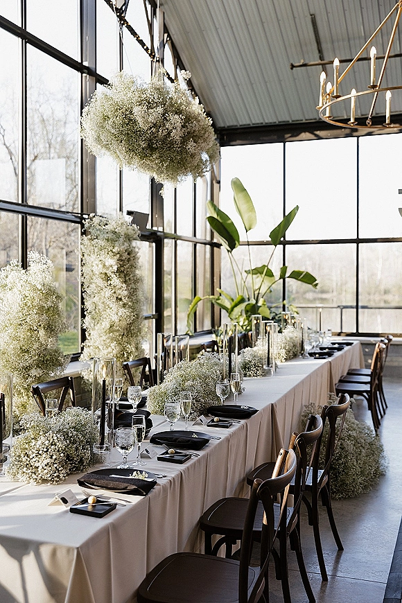 Reception tablescape with long banquet table setting, baby’s breath garland and hanging flowers, floating candles by sunlit glass walls