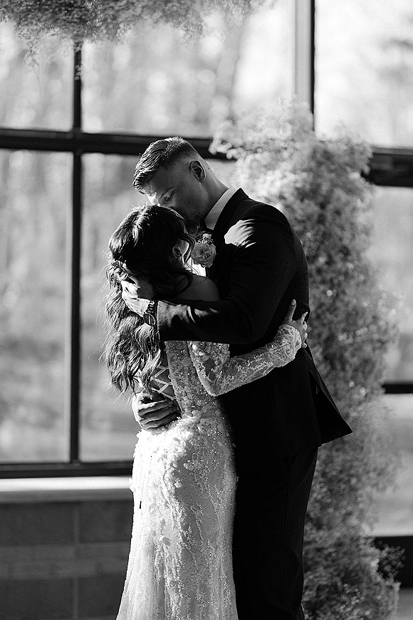 Wedding kiss portrait of bride and groom kissing by a large window in natural light, her lace long-sleeve dress framed by floral backdrop