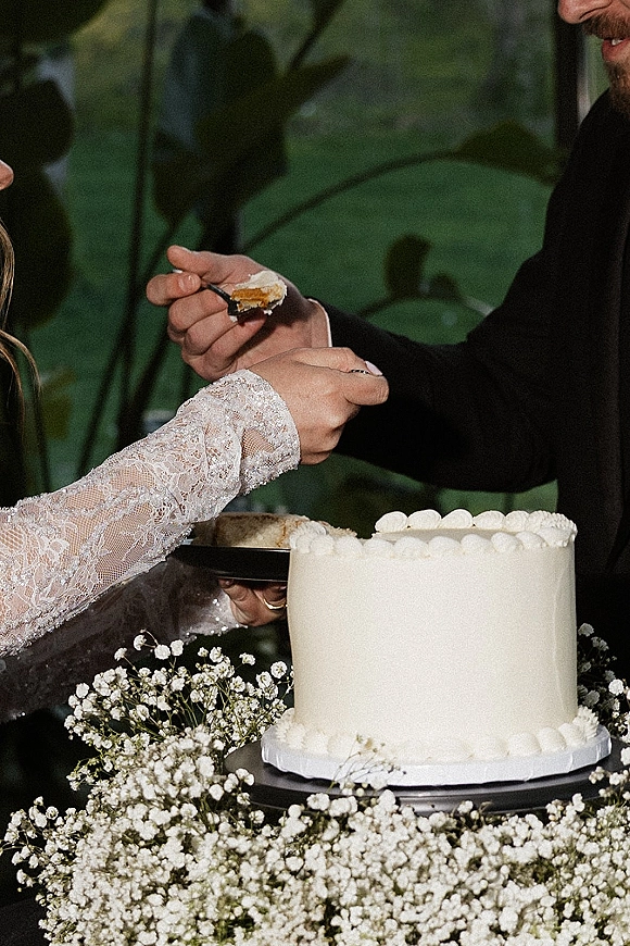 Cake cutting as bride in lace sleeve and groom in black suit slice white frosted wedding cake on stand in a greenhouse setting