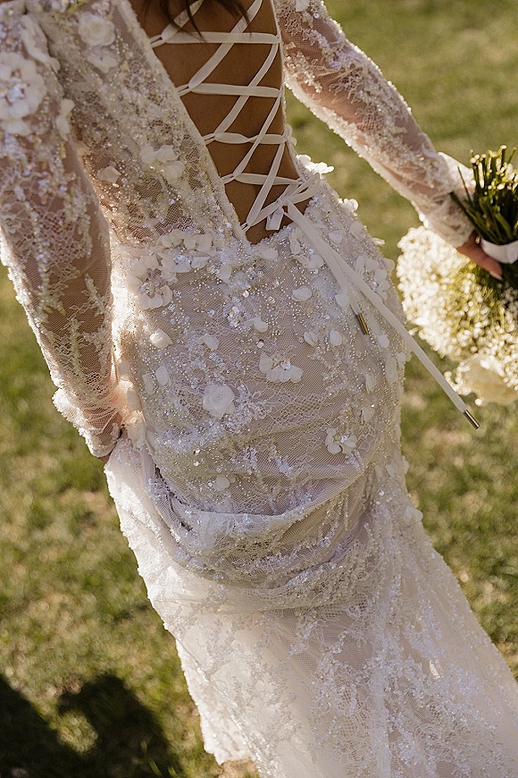 Wedding dress close-up of an open back wedding dress with corset lacing, lace sleeves and beaded 3D florals, bouquet on grass lawn