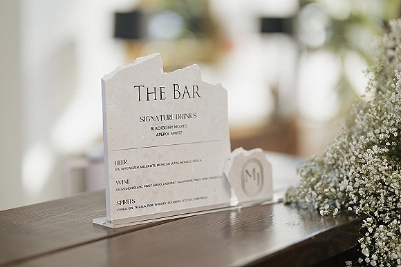 Wedding bar sign with signature drinks sign text on clear acrylic, framed by baby's breath on a bar counter with warm bokeh lights