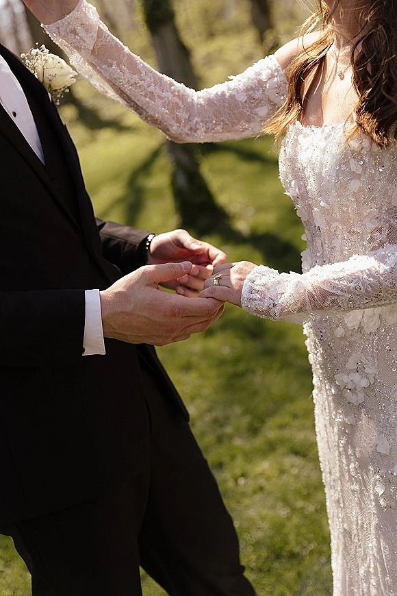 Wedding ceremony moment as groom places a ring on the bride’s finger, their hands framed by lace sleeves and black tuxedo on a tree-lined lawn
