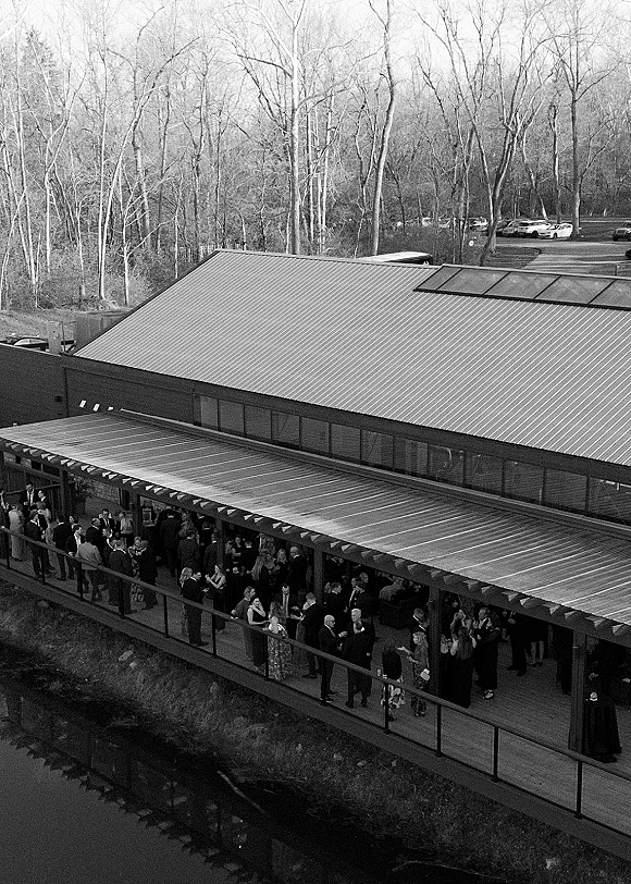 Wedding cocktail hour guests mingle with drinks around high-top tables on a covered patio with railing, creek and trees beyond