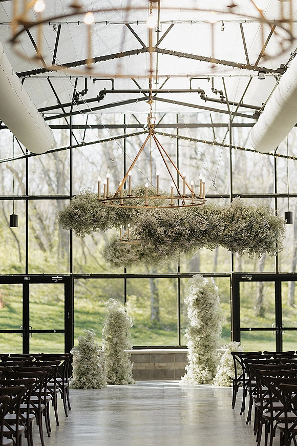 Indoor ceremony setup in a greenhouse wedding ceremony with a chandelier of baby's breath, taper candles, and floral pillars under string lights