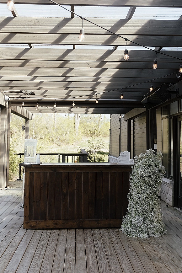 Wedding bar setup on a wood bar with string lights and floral arrangement, featuring a bar sign and drink dispenser on a covered patio deck
