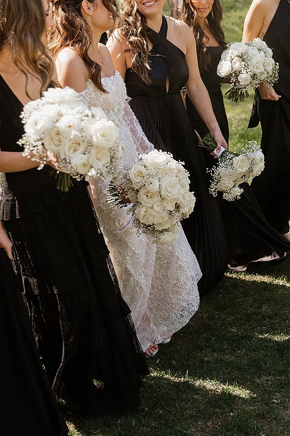 Bridesmaids with bride in black bridesmaid dresses holding white rose and baby’s breath bouquets on a green lawn outdoors