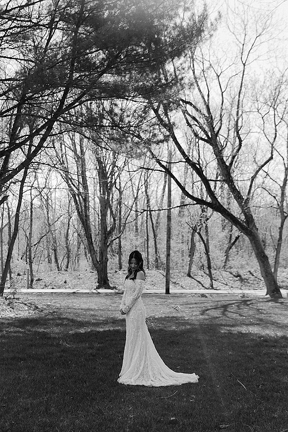 Bridal portrait of a bride looking down in an off-the-shoulder lace gown with a long train, standing on a wooded park pathway