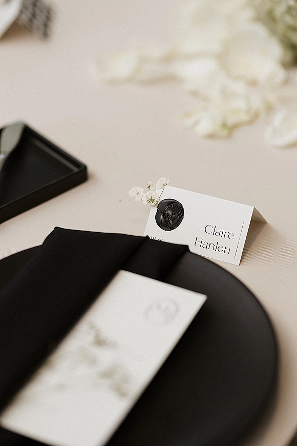 Wedding place card with a black wax seal and small white flowers, tied with a black ribbon on a light linen tablecloth backdrop